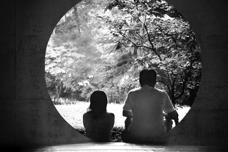 Two silhoetted people sit in front of a circular doorway, looking at green trees