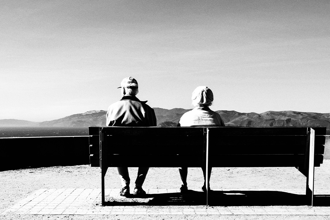 Ältere Frau und älterer Mann sitzen auf einer Bank und schauen in die Ferne.
