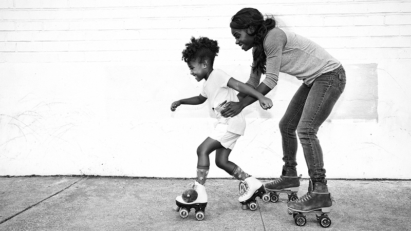Adult helping a child learn to roller skate on a sidewalk, both laughing and moving forward together.
