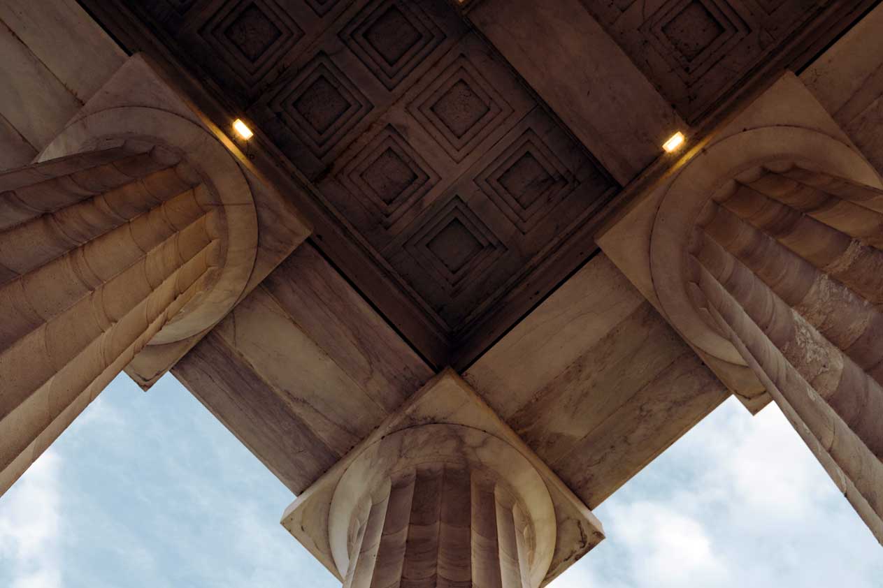 Vista dal basso di colonne classiche in pietra che si incontrano in un angolo, con dettagli decorativi del soffitto e luce soffusa sopra.