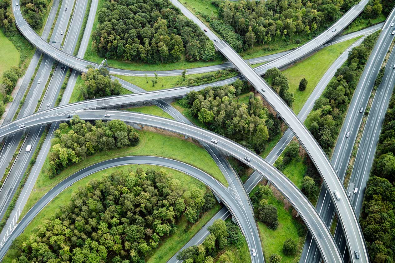 Luchtbeeld van een complex snelwegknooppunt met meerdere viaducten die door groene, met bomen gevulde omgeving kronkelen en licht verkeer.