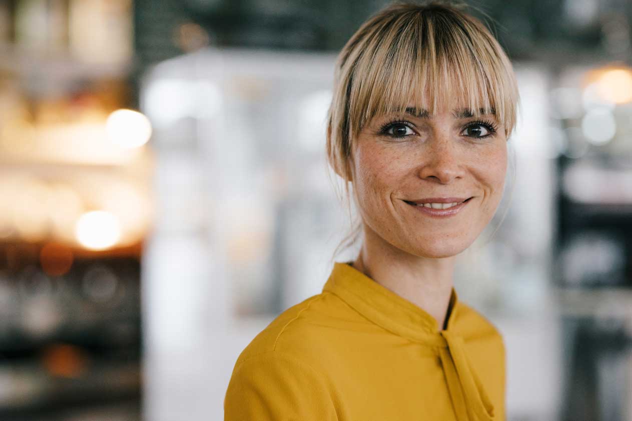 Primo piano di una donna sorridente con capelli biondi e frangia, che indossa un top giallo al chiuso.
