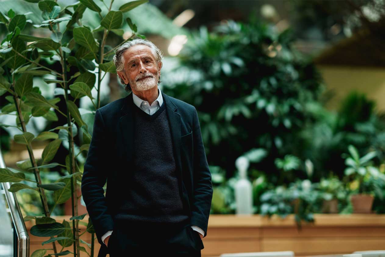 Oudere man staand in een lichte binnenruimte met planten, handen in de zakken, rustig kijkend naar de camera.
