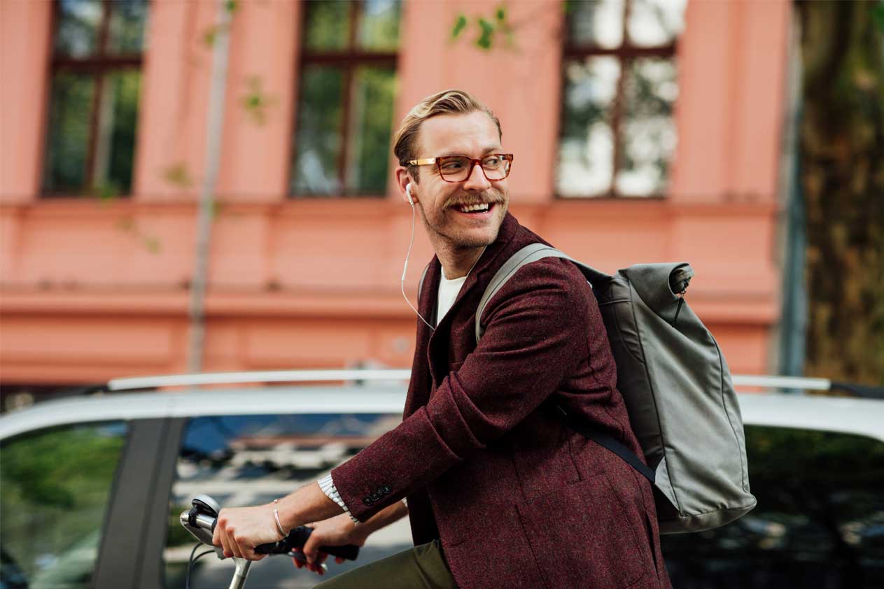 Uomo che va in bicicletta con uno zaino, sorridendo e guardando di lato su una strada cittadina.