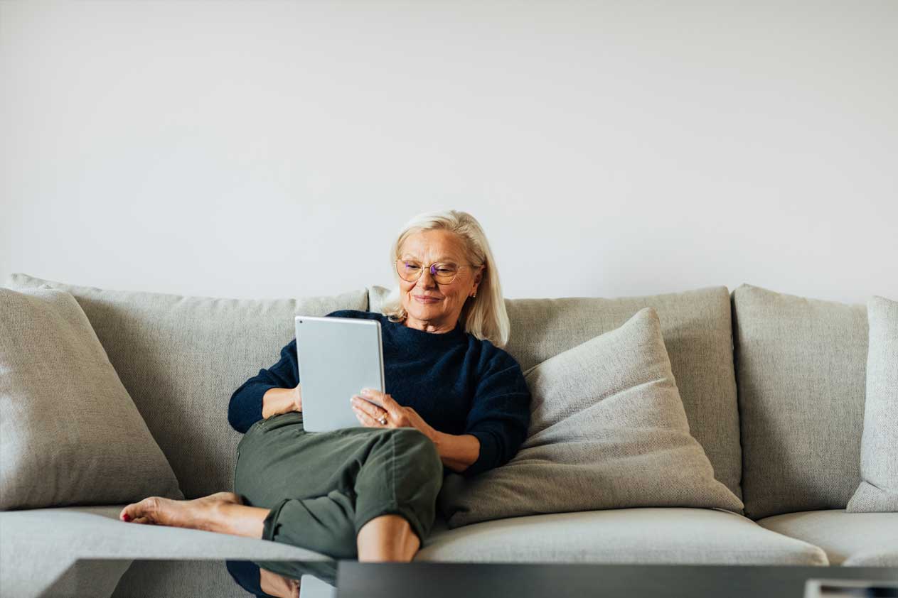 Donna anziana seduta su un divano a casa che usa un tablet, con cuscini e un ambiente interno neutro.