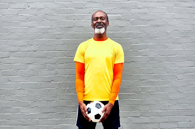  Mature man with a grey beard wearing a bright yellow and orange sports top, holding a black-and-white football in front of a grey brick wall.