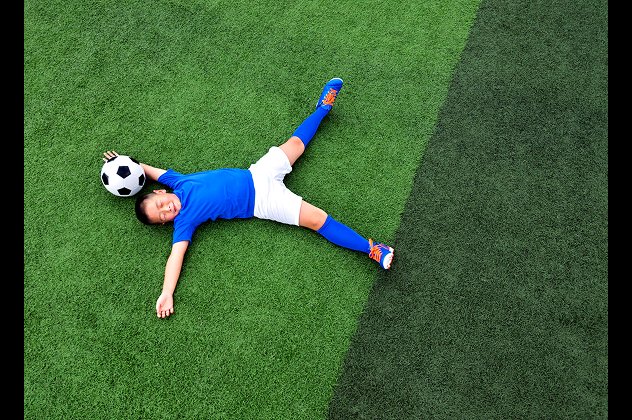 Young soccer player in a blue kit lying on a green grass football pitch with a soccer ball resting under his head.