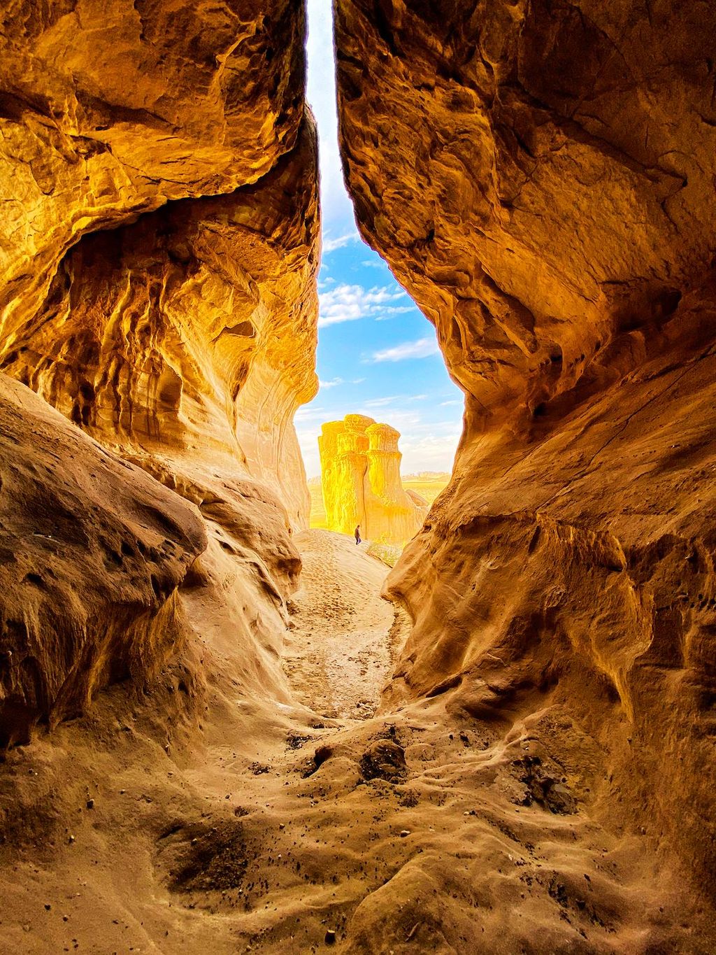 View through a narrow sandstone canyon opening toward a sunlit rock formation in a desert landscape.