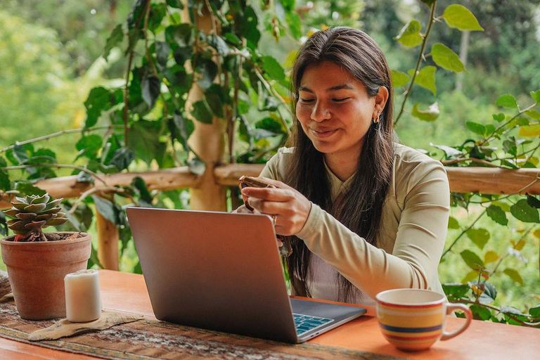 Person working on a laptop at an outdoor table surrounded by plants, holding a smartphone beside a coffee mug.