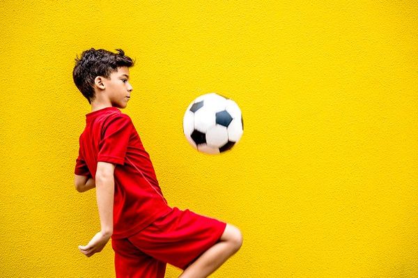 Young boy in a red football kit practising keepy-uppy skills with a black-and-white soccer ball against a bright yellow wall.