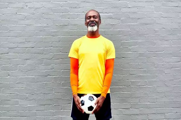 Mature man with a grey beard wearing a bright yellow and orange sports top, holding a black-and-white football in front of a grey brick wall.
