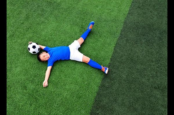 Young soccer player in a blue kit lying on a green grass football pitch with a soccer ball resting under his head.